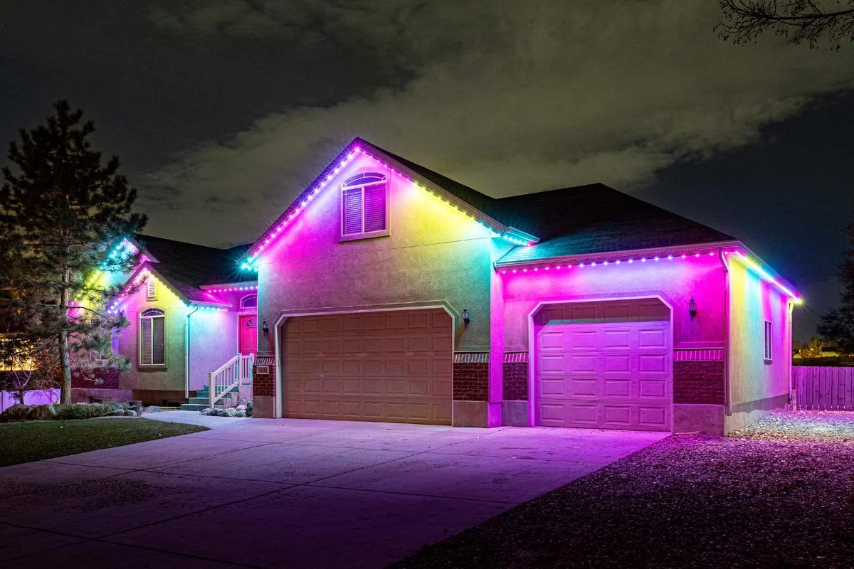 37-1 A single-story house in New Franklin at night is illuminated with vibrant pink, purple, and blue permanent lighting outlining the roof and garage, creating a colorful and festive appearance.