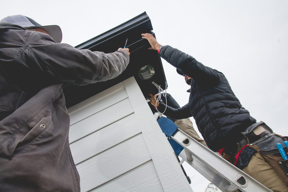 Two people work together on a ladder, handling holiday lighting installation near the roof of a white building in New Franklin. Tools and cables are visible as they work beneath an overcast sky.