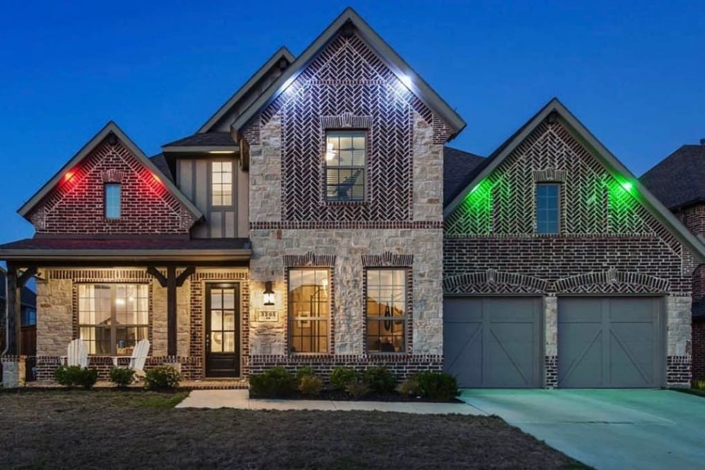 4 A two-story brick house with a porch, stone accents, two garage doors, and colorful exterior lights glowing red, white, and green at dusk. Two chairs sit on the front porch near the entrance.