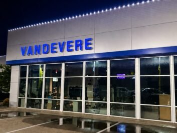 1 A car dealership at night with VANDEVERE in blue letters above large glass windows. Reflections and empty parking spaces are visible in the wet pavement outside.