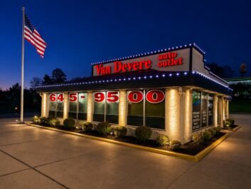 11 A brightly lit Van Devere auto outlet building at night, with illuminated red signage and an American flag on a pole outside. Large numbers are displayed on the windows.