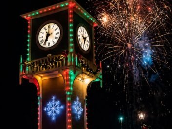 5 A clock tower with colorful holiday lights and glowing snowflakes stands at night, as fireworks burst in the sky. A crowd gathers below, with festive street decorations visible in the background.