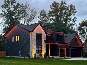 8 Modern two-story house with dark siding, stone accents, and a double garage. Warm lights outline the roof, and large windows glow at dusk. Surrounded by grass and tall trees.