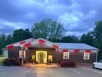 9 A single-story gray building with a metal roof, lit by red and white lights along the eaves. Two rocking chairs sit by the front door. Trees and a cloudy sky are in the background.