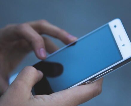 A person holds a white smartphone with both hands, touching the screen with their right index finger. The phones screen is off or locked, reflecting a faint blue tint. The background is out of focus.